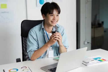 Neşeli genç bir profesyonel elinde bir kahve fincanı tutarken ve bir masada dizüstü bilgisayarla otururken gülümserken, pozitif bir çalışma ortamını somutlaştırıyor..