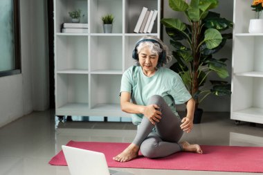 Son sınıf öğrencisi bir kadın evde sağlık egzersizlerine katılıyor, kulaklık takıyor ve yoga minderinde otururken internetten dersini takip ediyor..