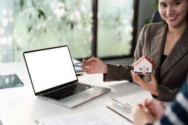 Bir emlakçı müşterisiyle mülkiyet seçeneklerini büyük bir hevesle tartışır. Model bir evi şık bir ofis alanında görsel yardım olarak kullanır..