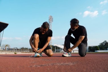 İki kıdemli adam ayakkabı bağcıklarını piste bağlayıp takım çalışmasını ve sağlık yolculuğunun önemini vurguluyor..