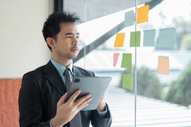 Odaklanmış bir profesyonel inceleme, stratejik planlamayı vurgulayarak, elinde bir tablet tutarken yapışkan notlar üzerine proje ayrıntılarını inceler.