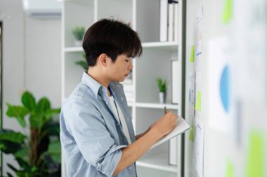 Genç bir iş analisti, çeşitli veri tablolarını incelerken notlar alır, analitik becerileri ve modern ofis ortamındaki detaylara dikkat eder.