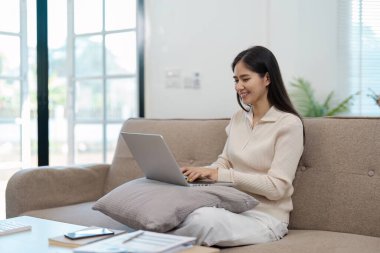 Neşeli, genç Asyalı bir kadın koltuğuna oturmuş laptopuna odaklanmış, verimli ve neşeli bir çalışma alanını somutlaştırıyor..