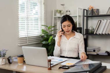 Parlak bir ofiste, bir kadın önemli belgeleri incelerken mali konuları tartışmak için bir telefon görüşmesi yapar..