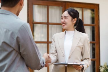 Gülümseyen bir emlakçı, müşterisiyle evin dışında tokalaşırken, satın alımın başarıyla tamamlanması için arkadaşça ve profesyonelce bir hava katıyor..