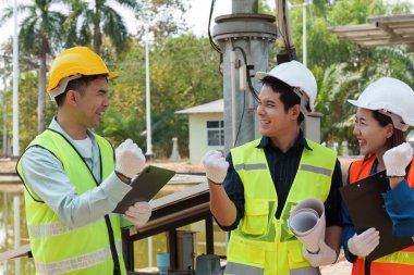 Sert şapka ve güvenlik teçhizatıyla donatılmış üç inşaat çalışanı, bölgede zafer anı paylaşıyor ve sürdürülebilir uygulamaların uygulanmasında coşku ve işbirliğini yansıtıyor