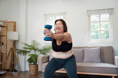 Neşeli, iri yarı bir kadın evde egzersiz yapıyor, halterleri kaldırırken gülümsüyor, vücut pozitifliğini ve sağlığını arttırıyor..