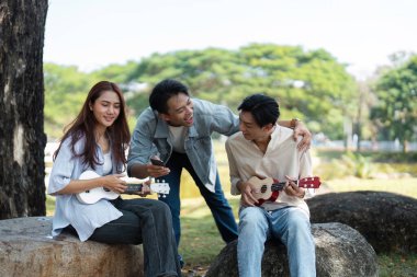 Bir grup arkadaş dışarıda dinleniyor, müzik ve kahkahaları dingin bir parkta ukulelelerle paylaşıyorlar.