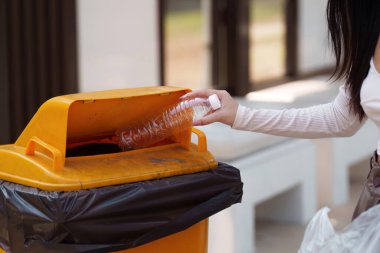 Bir kişi, geri dönüşüm ve çevre bakımının önemini vurgulayarak, sarı bir geri dönüşüm kutusuna plastik bir şişe koyar..