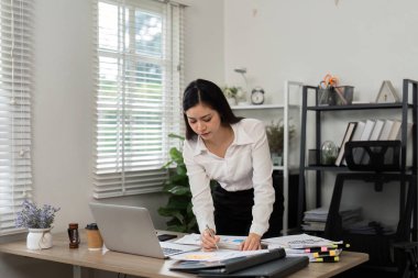 Masasının başında durmuş belgeleri inceleyen odaklanmış bir işkadını, mali bir ortamda çalışkanlık ve profesyonelliği vurguluyor.