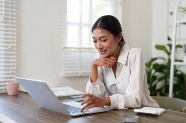 Bu görüntü, bilgisayarında özenle çalışan bir iş kadınını gösteriyor. Uzak çalışma ortamında hedeflerine ulaşmak için teknolojinin verimliliğini ve etkili kullanımını vurguluyor..