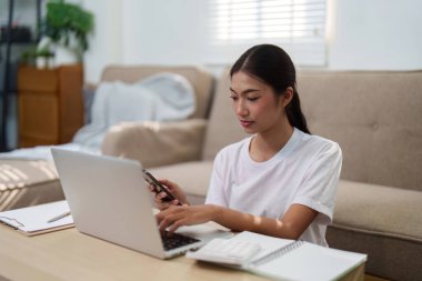 Dizüstü bilgisayarı ve akıllı telefonunu kullanan odaklanmış genç bir kadın, uzaktan çalışmada gerekli nişan ve verimliliği vurguluyor.