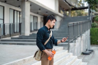 Genç bir adam açık hava basamaklarında duruyor, elinde parlak turuncu bir su şişesi tutarken, teknolojinin ve sürdürülebilir alışkanlıkların bir karışımını gösteriyor..