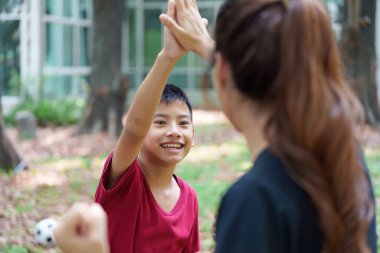 Genç bir çocuk parkta bir kadına beşlik çakarken mutlulukla ışıldıyor. Bu, spor yaparken cesaret ve aile bağını simgeliyor..