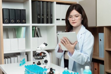 Modern bir laboratuvarda, laboratuvar önlüğü ve gözlükleri giymiş bir araştırmacı bir tablet üzerindeki araştırma verilerini inceleyerek, teknolojinin bilimsel çalışmalara entegrasyonunu vurguluyor..