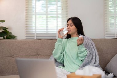 Kanepesinde rahat bir şekilde, genç bir kadın su içiyor ve ilaç alıyor, bir anlık kendine bakımı somutlaştırıyor ve huzurlu bir ev ortamında sağlığını büyütüyor..