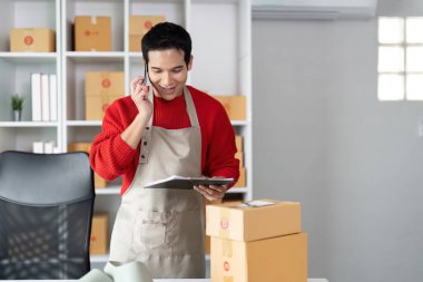 Adanmış bir girişimci, ambalaj emirleri arasında, e-ticarette gereken özeni yansıtarak, nakliye detaylarını telefonda koordine ederken bir tablet hakkında ayrıntılı notlar alır..