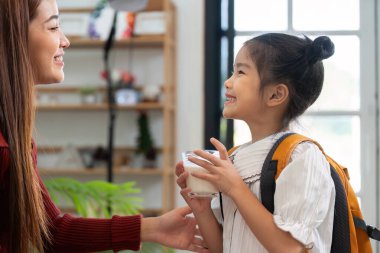 Annesi ona gülümserken, mutlu bir kız elinde bir bardak süt tutuyor, okul günü için heyecanlanıyor. Bu an, okula gitmeden önce beslenme ve sabah rutinlerinin önemini gösterir..