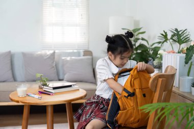 Küçük bir kız masada parlak turuncu çantasını karıştırıyor ve okul organizasyonunun heyecanını sergiliyor..