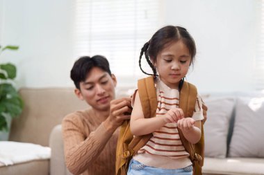 Sevgi dolu bir baba, kızına sırt çantasını takmasında yardımcı olur. Onu okula hazır ve kendinden emin hissettirir. Sahne, ebeveyn desteğinin desteklendiğini vurguluyor.