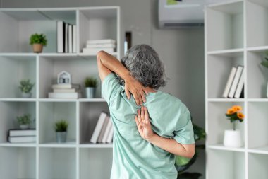 Yaşlı kadın kollarını arkasından uzatarak, huzurlu ev ortamında esneklik egzersizi yaparak, sağlıklı yaşam ve aktif yaşlanmayı teşvik ediyor..