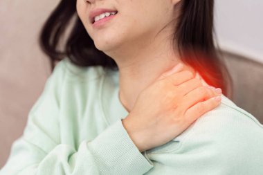 Yeşil kazaklı bir kadın omzunu tutarak acı ve rahatsızlığı, sağlık hizmetlerini ve tıbbi bakım ihtiyacını sembolize eder..