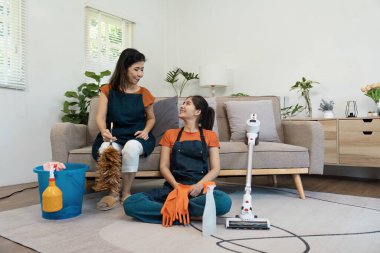 Önlüklü bir anne ve kız temizlik malzemeleri ve bitkilerle çevrili, aydınlık ve modern bir oturma odasında temizliğe ara veriyorlar..