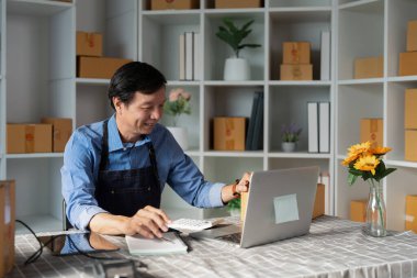 Bir Asyalı girişimci, bir dizüstü bilgisayarla e-ticaret faaliyetleri yaparken gülümserken, modern bir başlangıç ortamında pozitif bir atmosfer yaratıyor..