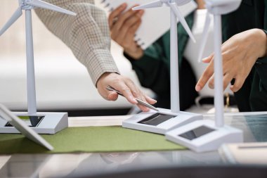 Bir iş ortamında rüzgar türbini modelleri tarafından temsil edilen yenilenebilir enerji çözümlerine odaklanarak sürdürülebilirlik uygulamaları hakkında tartışan iki kişi.