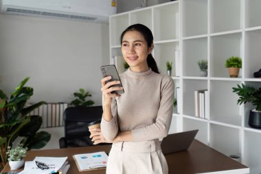 Şık bir ofis ortamında, elinde akıllı telefon ve kahve, modern iş ve teknolojiyi somutlaştıran genç profesyonel bir kadın..