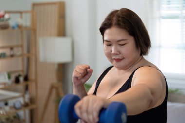 Kilolu bir kadın evinde dambıllarla antrenman yapıyor. Rahat bir ortamda spor yapmaya ve kendini geliştirmeye kendini adamış..