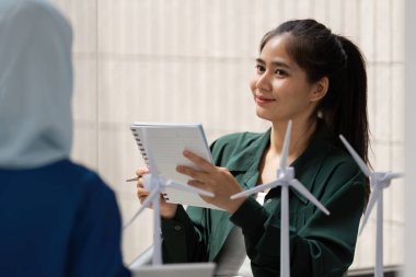 Odaklanmış bir kadın, çevre dostu enerji çözümlerini sembolize eden rüzgar türbini modelleriyle çevrili sürdürülebilirlik girişimleriyle ilgili bir toplantıda notlar alırken gülümsüyor..