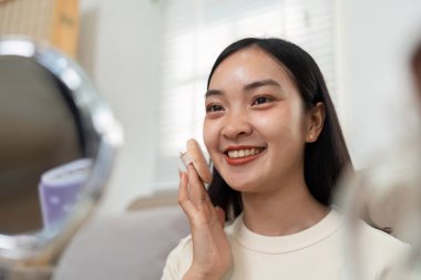 Parlak bir mekanda, genç bir kadın cilt rutininden zevk alır, mutluluk ve güç saçan bir gülümseme ile bir ürün uygular..