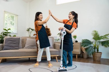 İki mutlu hizmetçi aydınlık, modern bir oturma odasını temizledikten sonra, takım çalışması ve ev işinden memnuniyet göstererek beşlik çakar..