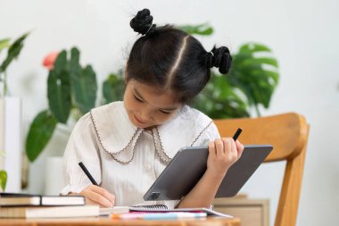 Küçük bir kız ödevine özenle odaklanıyor, tablet ve not defterini dengeliyor, modern eğitim ve yaratıcılığın ruhunu somutlaştırıyor..