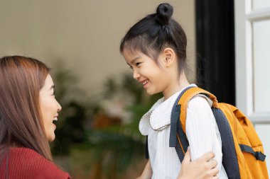 Bir annenin kızını kucakladığı, okula hazırlanırken sevgisini ve desteğini sergilediği neşeli bir an. Sırt çantası takan küçük kız, coşkuyla ışıldıyor..