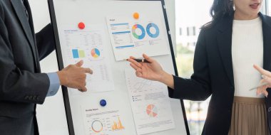 Stratejik bir toplantı alanında, ekip üyeleri bütçe raporlarını tartışırken, bir kişi tahtadaki kritik verilere işaret ediyor. Nişan ve etkileşim gelişir.