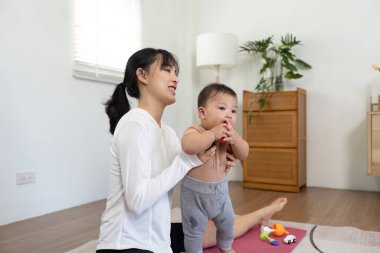 Bir anne, ayakta durmayı öğrenirken bebeğine destek olur. Çocuk gelişimi ve oyun oynamanın heyecan verici yolculuğunu vurgular..