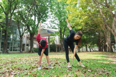 Enerjik bir çocuk ve bir kadın birlikte esneme egzersizleri yapıyor, yemyeşil bir parkta birbirlerine eşlik ederken fiziksel kondisyonlarını geliştiriyorlar..