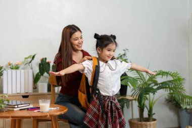 Sırt çantası takan genç bir kız, sevinçle poz verirken annesi yeni bir okul yılına hazırlanmasına yardım ediyor, heyecanlı bir anı yakalıyor..