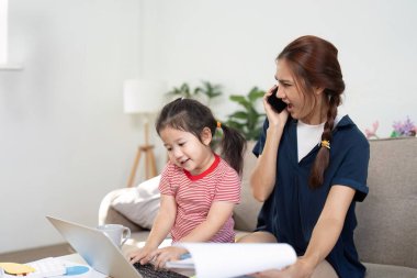 Çoklu görev yapan bir anne, kızına dizüstü bilgisayarda yardım ederken bir telefon görüşmesi yönetir ve modern bir ev ortamında ebeveynliğin zorluklarını ve zevklerini anlatır..