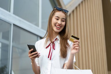 Okul kıyafeti giymiş mutlu bir öğrenci, elinde kredi kartı ve alışveriş çantası tutarken gülümsüyor, okula dönüş alışverişinin heyecanını yakalıyor..