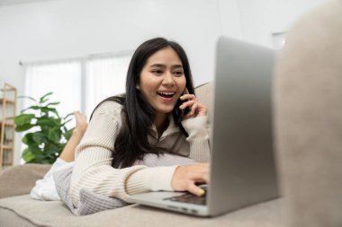 Neşeli bir kadın kanepesinde uzanıyor, bilgisayarına erişirken canlı bir telefon görüşmesi yapıyor, modern iş hayatında konfor ve bağlantının karışımını gösteriyor..