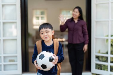 Okuldaki ilk günü için heyecanlanan genç bir çocuk, annesi el sallarken bir futbol topu tutuyor. Bu resim çocukluk sevincini ve aile teşvikinin önemini vurgular..