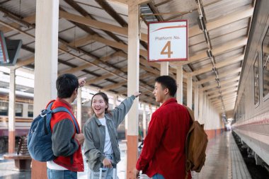 Hevesli arkadaşlar platformun tabelasına bakar, onu gösterir ve bir sonraki seyahat noktalarını tartışır, keşif ve macera ruhunu somutlaştırır..