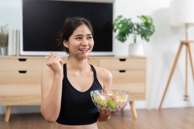 Neşeli bir yüzü olan genç bir kadın taze salatanın tadını çıkarıyor, sıhhatli bir yaşam tarzını ev ortamında yansıtıyor..