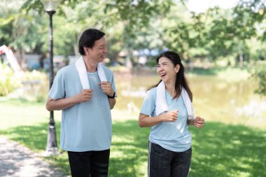 Aktif kıdemli bir çift yemyeşil bir parkta birlikte yürür, sağlık yolculuklarına odaklanırken gülücüklerini ve kahkahalarını paylaşırlar. Aralarındaki bağ fiziksel aktivitenin önemini vurguluyor..