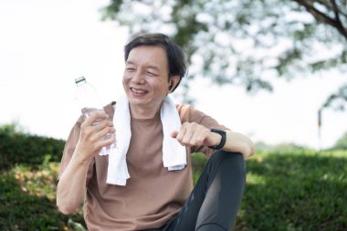 Çimlerin üzerinde oturan neşeli, kıdemli bir adam, yoga seansından sonra suyun tadını çıkarırken geniş bir gülümseme, aktif yaşam ve refahı gösteriyor..