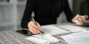 Odaklanmış bir iş adamı finansal belgeleri incelerken bir not defterine not alır, yardım için akıllı telefonu kullanır.