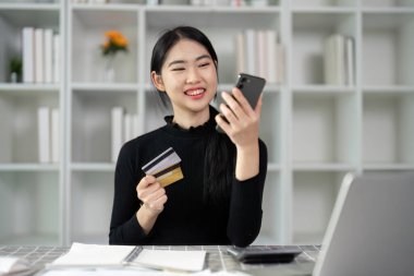 Neşeli genç bir kadın elinde bir kredi kartı tutarken akıllı telefonuna bakar ve internetten satın almaya hazırdır..